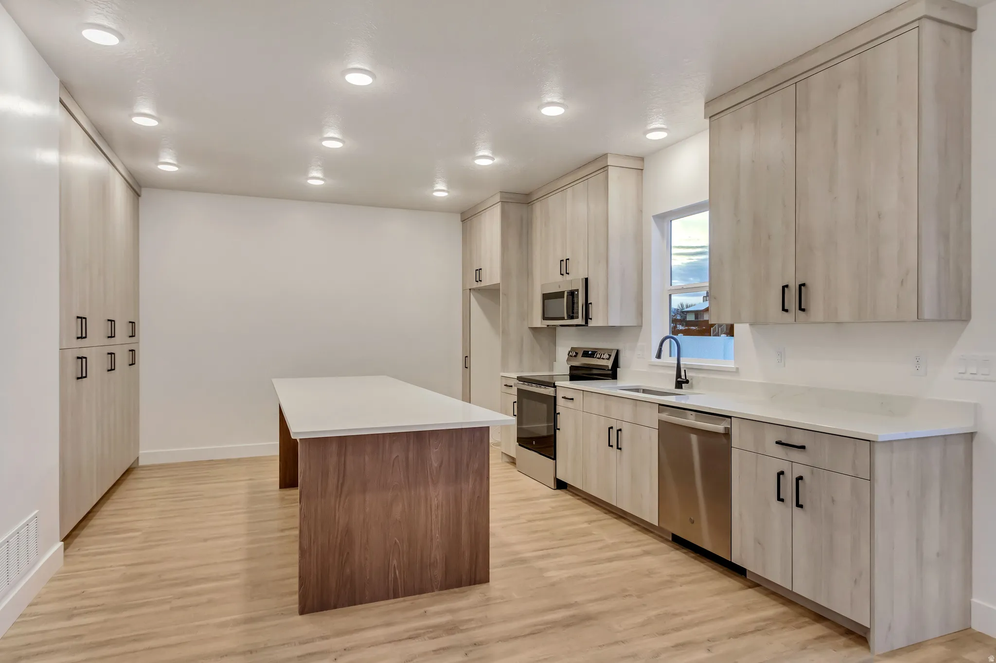Kitchen featuring an elongated island with a contrasting wood-finish base and light countertop, light wood-finish cabinetry, stainless steel appliances, and wood-finish flooring