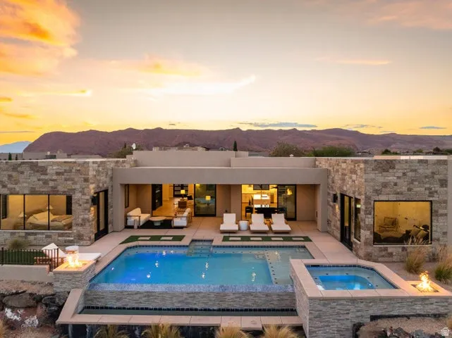 Back of house at dusk with an in-ground hot tub, a mountain view, an outdoor pool, a patio area, and an outdoor fire pit