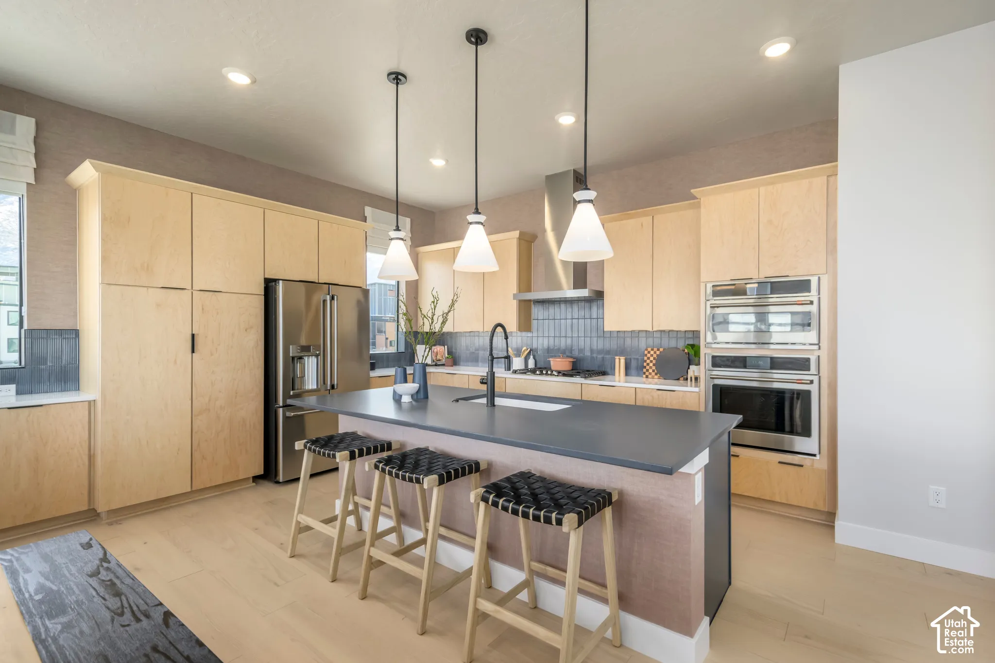 Kitchen featuring flat-panel wood cabinetry, a central island with a solid-surface countertop, stainless steel double wall ovens, a tiled backsplash, and three pendant light fixtures