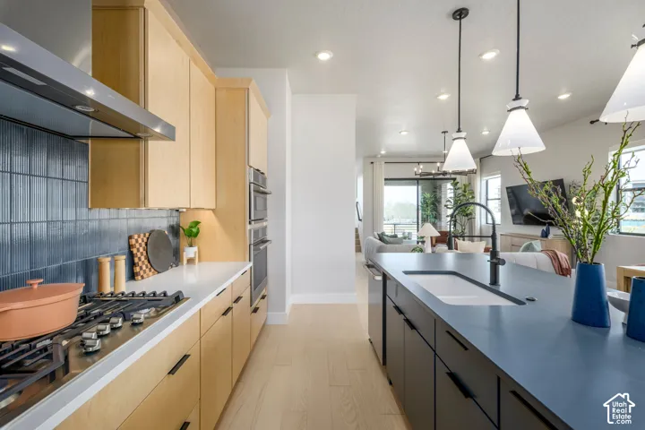 Kitchen featuring light wood-finish cabinetry, dark solid-surface countertops, blue vertical-tiled backsplash, stainless steel gas cooktop, and large central island with integrated basin and matte black fixtures