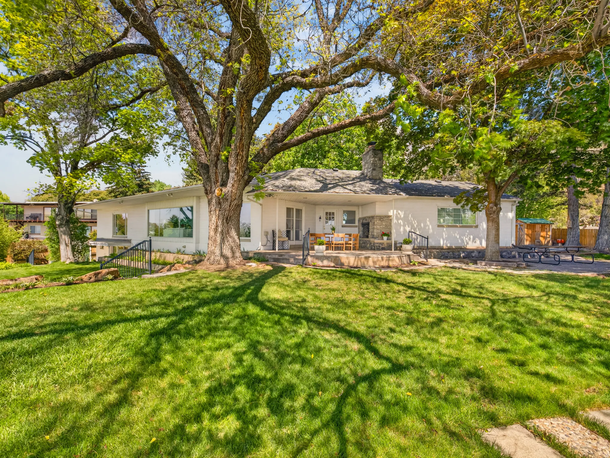 Expansive front lawn with mature trees and a stone patio featuring an outdoor fireplace