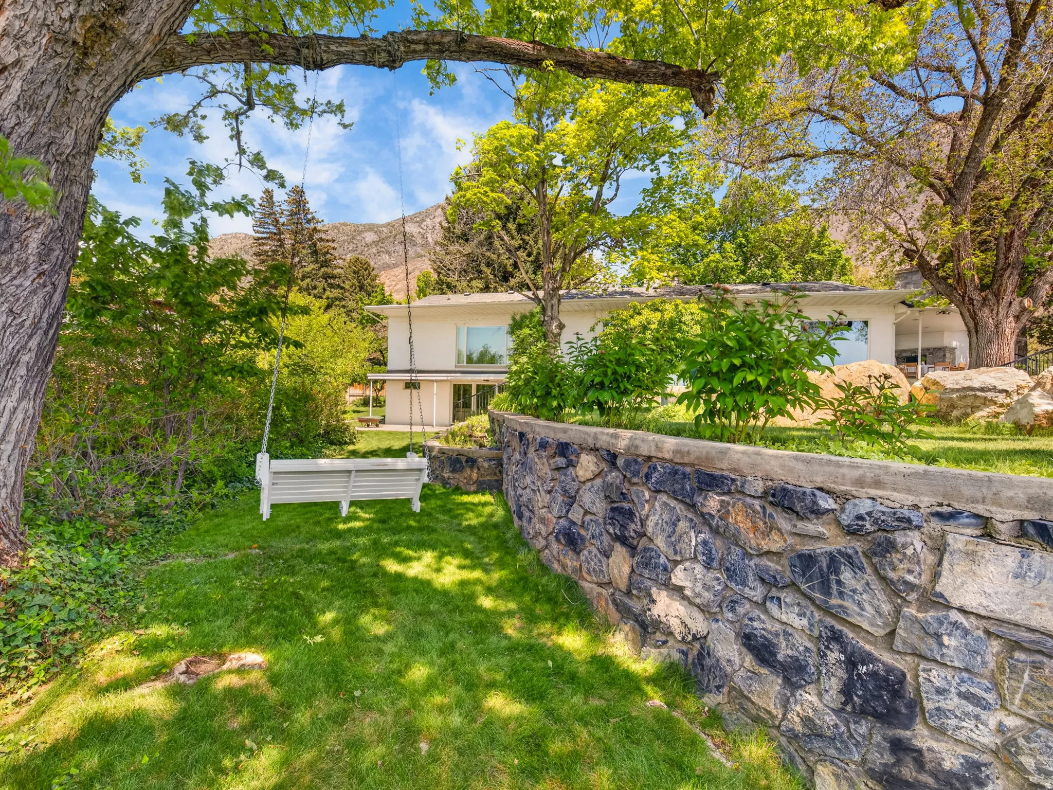 Expansive grassy yard featuring mature trees, a stone retaining wall, and a mountain backdrop