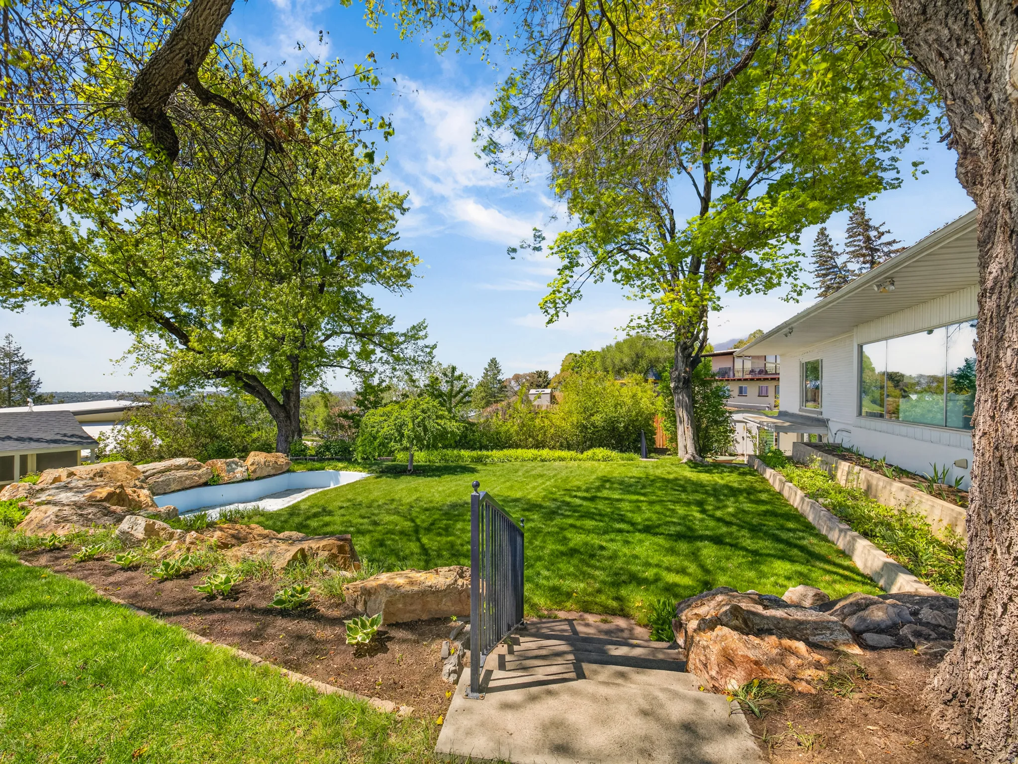 Expansive backyard featuring a lush lawn, established trees, a rock garden, concrete steps with a metal railing, and a white exterior home with large windows