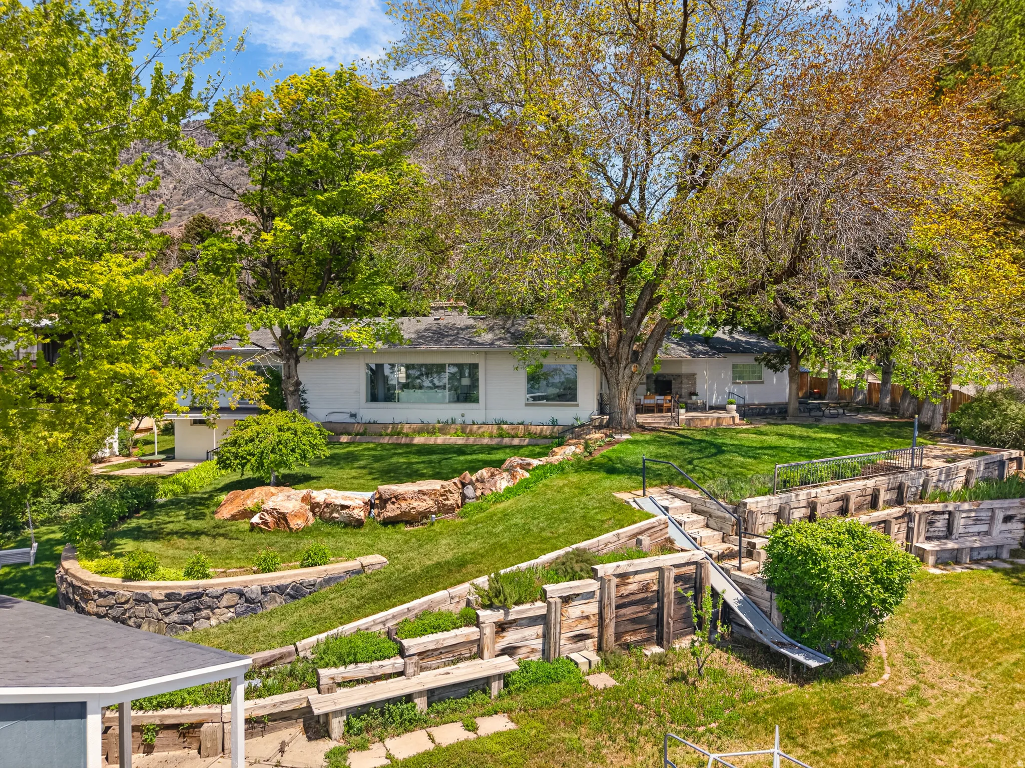 Expansive terraced backyard featuring a manicured lawn, stone retaining walls, and mature shade trees