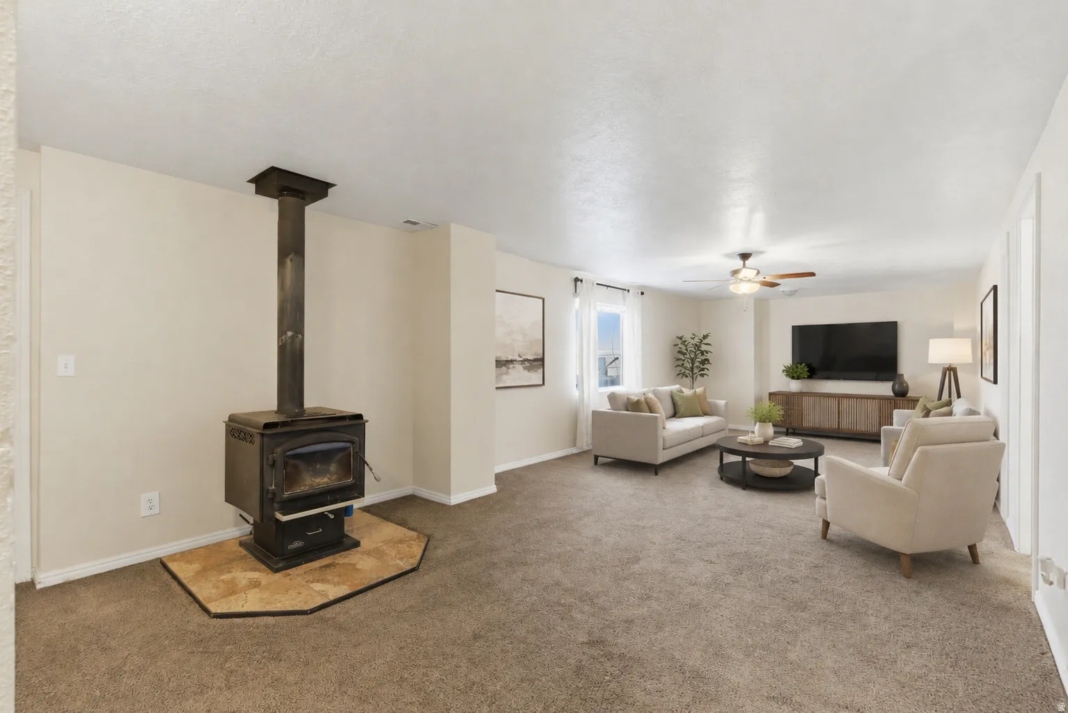 Spacious living area featuring a wood-burning stove with a tiled hearth, soft neutral carpeting, light-toned wall finishes, and a ceiling fan with integrated lighting