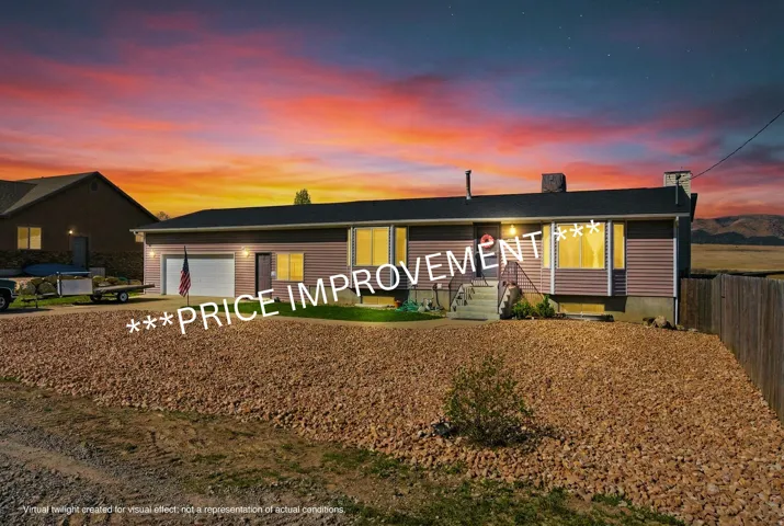 Modern ranch-style residence featuring a two-car garage, multiple illuminated windows with warm interior lighting, horizontal siding exterior, and a gravel front landscape with established plantings