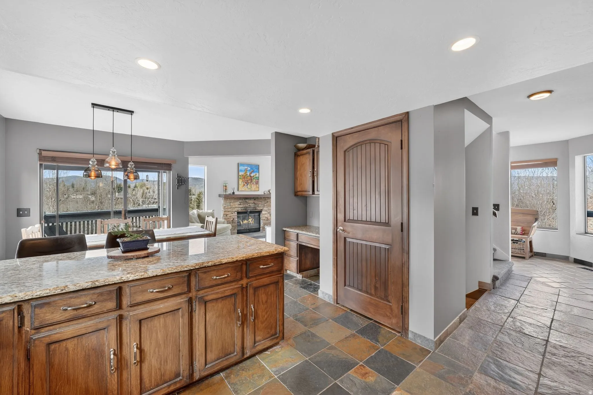 Kitchen featuring light stone counters, stone tile floors, a stone fireplace, wood finish cabinetry, and pendant lighting