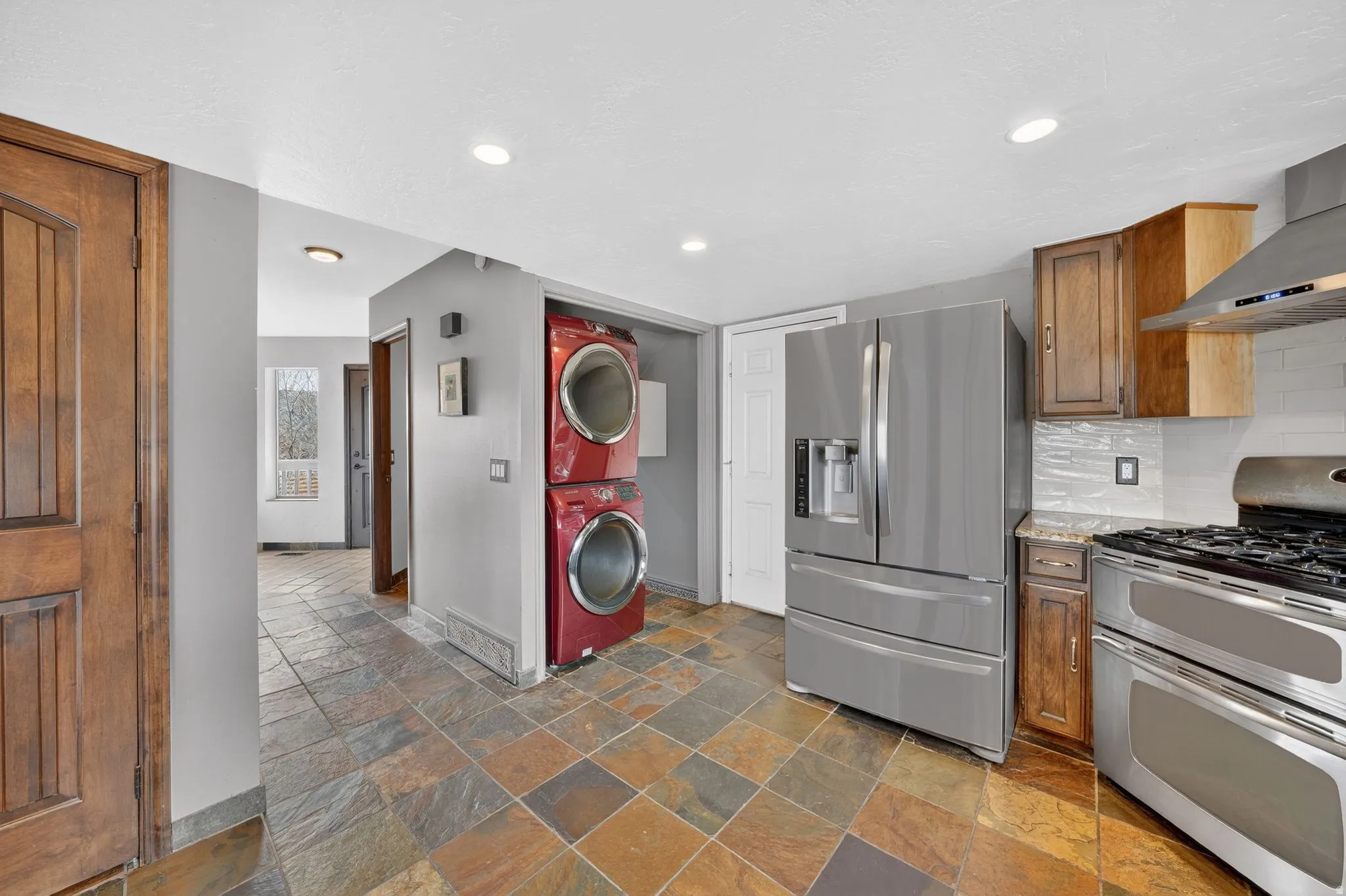 Kitchen featuring stainless steel appliances, wood finish cabinetry, stacked washing machine and dryer, light stone counters, and tasteful backsplash