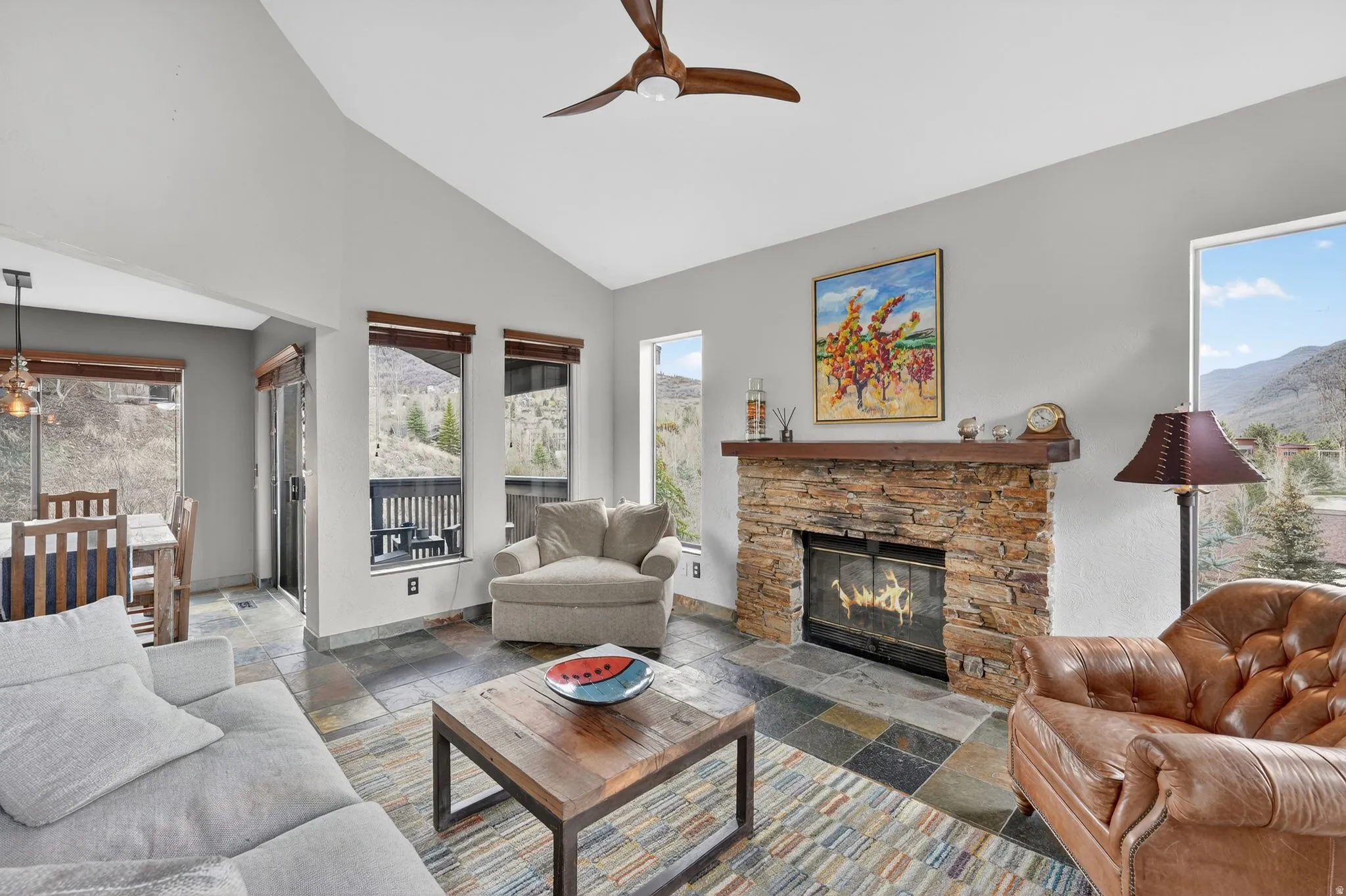 Living room with stone tile flooring, a fireplace, vaulted ceiling, ceiling fan, and healthy amount of natural light