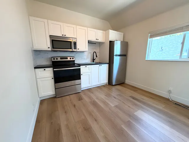 Kitchenette featuring stainless steel appliances, white cabinetry, dark countertops, a tiled backsplash, and wood-finish flooring