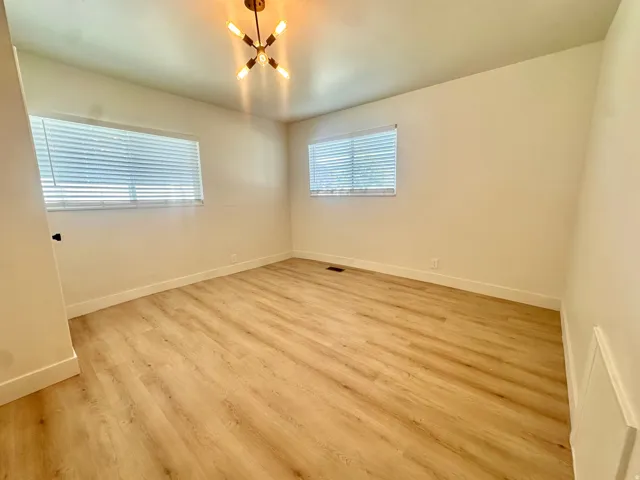Spacious room with wood-finish flooring, two windows with blinds, a modern ceiling light fixture, and white baseboards