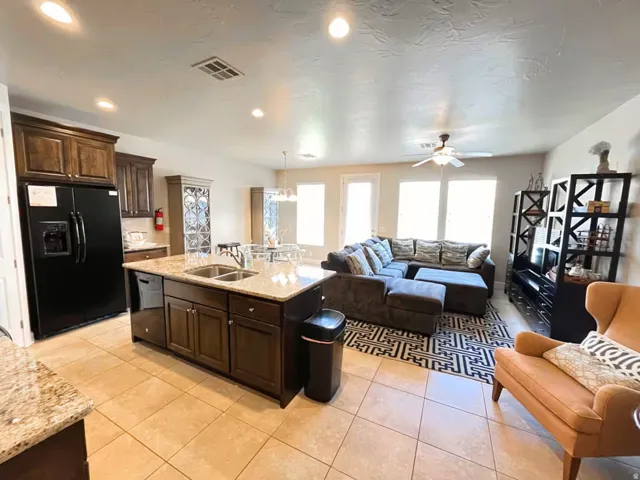 Kitchen featuring dark wood finish cabinetry, open floor plan, light stone counters, black appliances, and a center island with sink