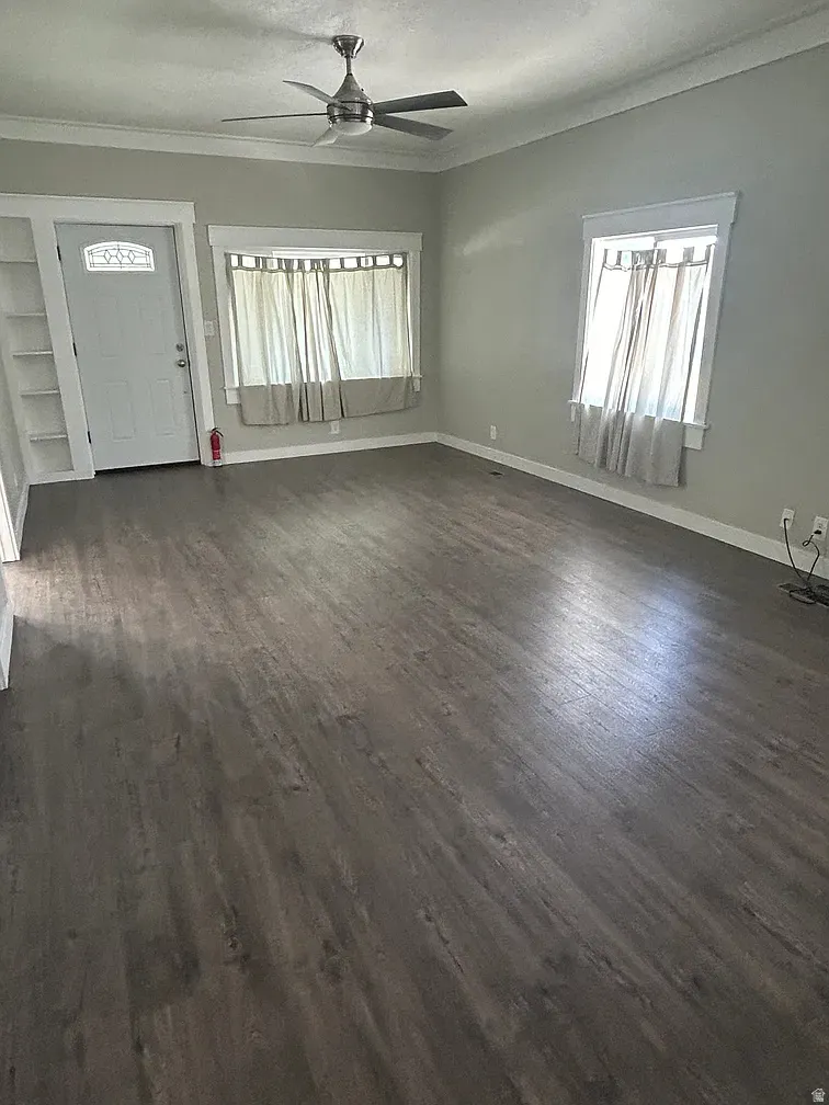 Unfurnished living room featuring dark wood-style floors, crown molding, ceiling fan, and plenty of natural light