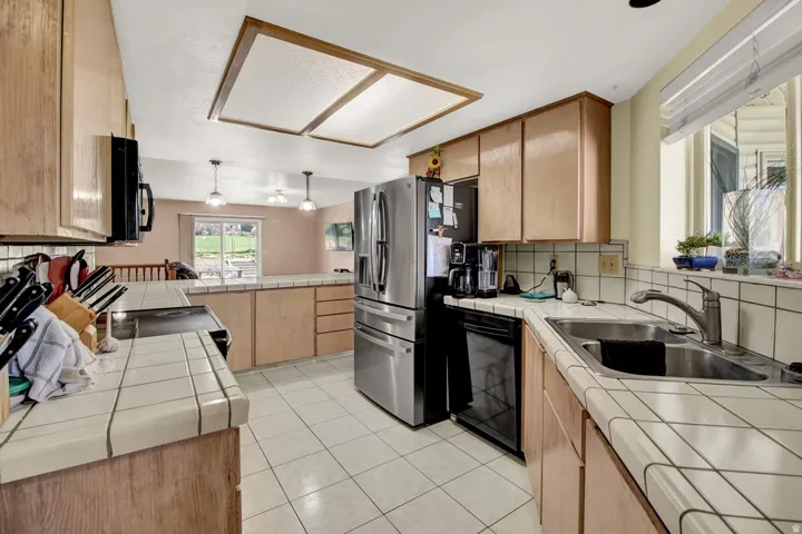 Kitchen with tile counters, light wood finish cabinetry, backsplash, a peninsula, and black appliances