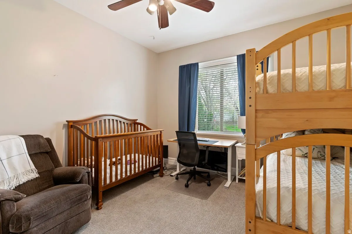 Bedroom featuring a crib, light colored carpet, an office area, and a ceiling fan