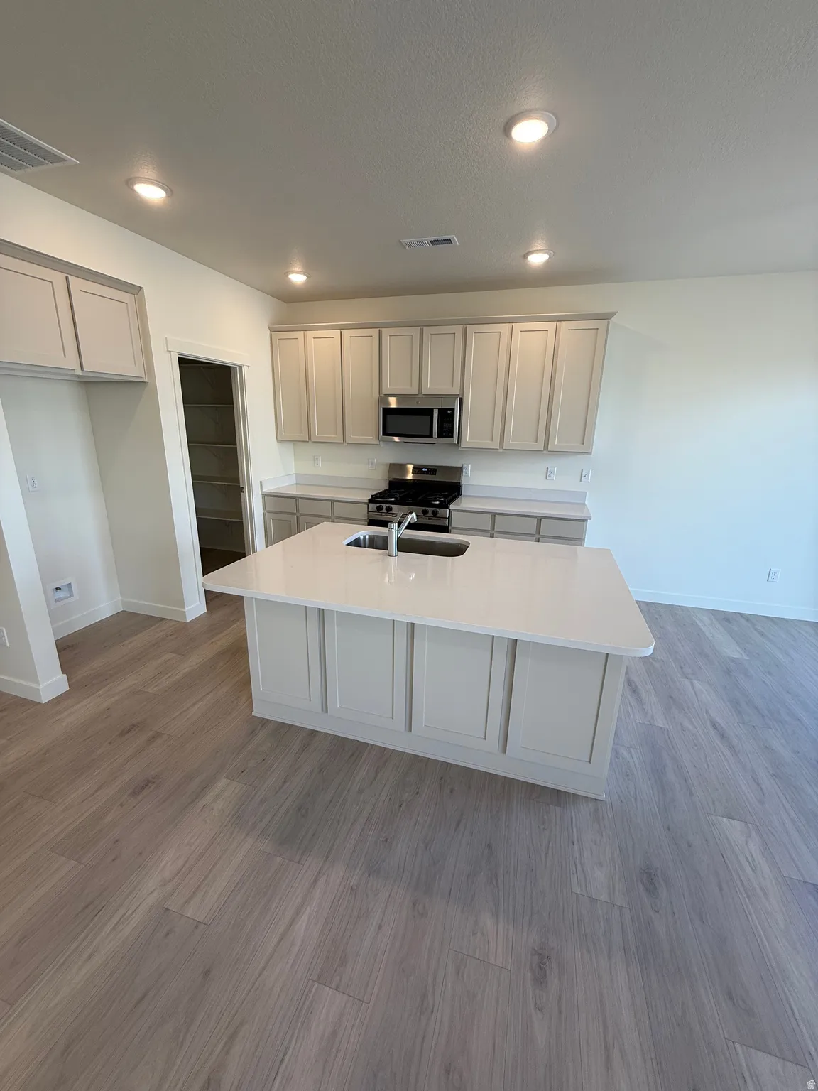 Kitchen featuring a central island with a sink, stainless steel appliances, light-colored cabinetry, and wood-finish flooring