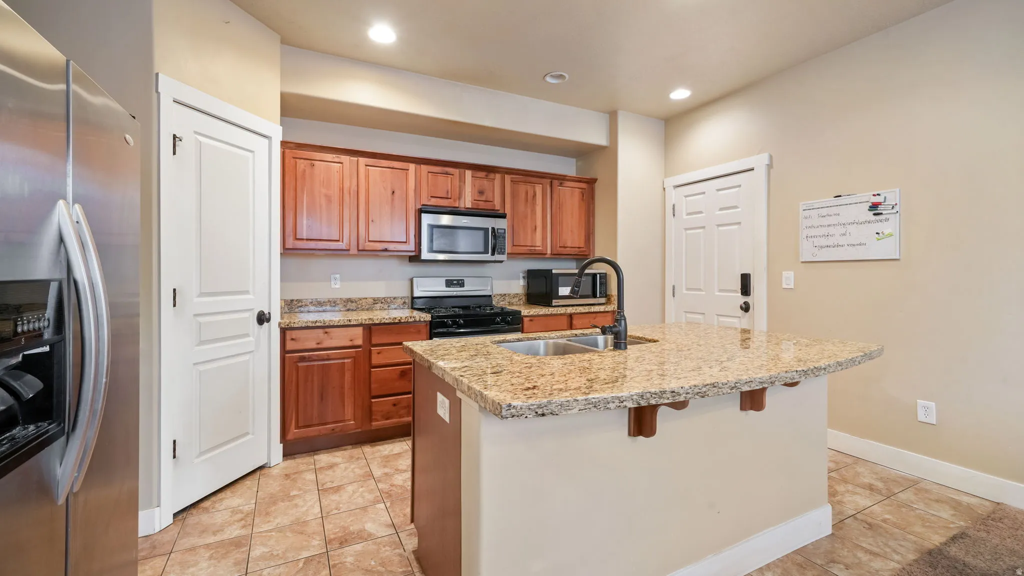 Kitchen featuring stainless steel appliances, wood finish cabinetry, light tile patterned flooring, light stone counters, and a center island with sink