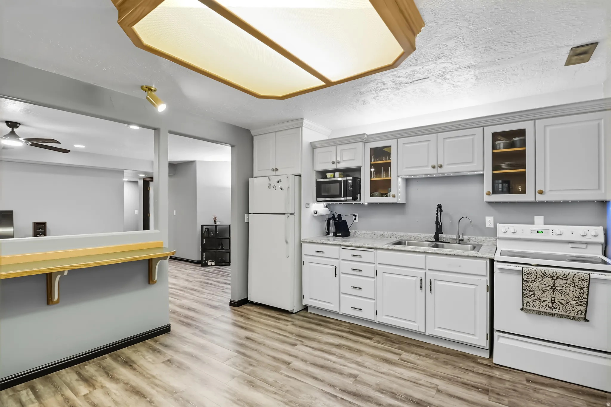 Basement kitchen featuring white appliances, light wood-type flooring, glass fronted cabinets, ceiling fan, and light stone countertops