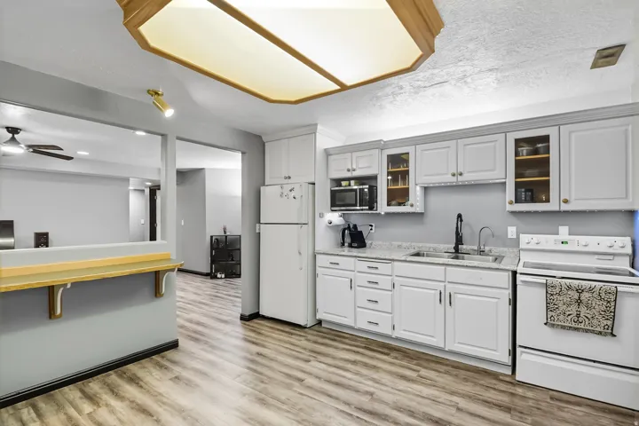 Basement kitchen featuring white appliances, light wood-type flooring, glass fronted cabinets, ceiling fan, and light stone countertops