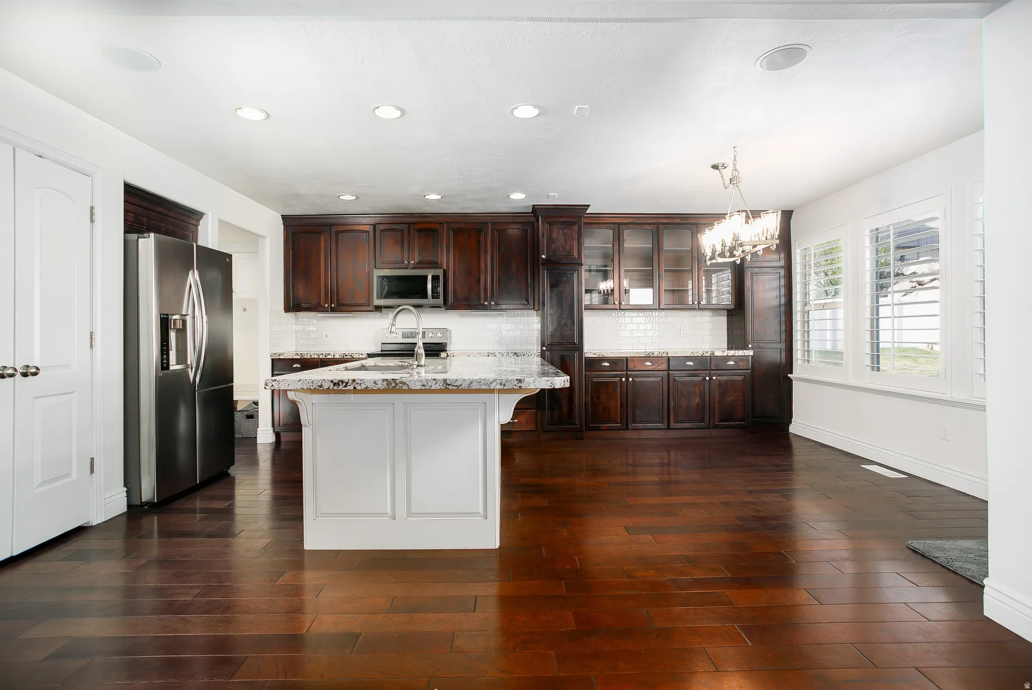 Kitchen with dark wood finish cabinetry, glass fronted cabinets, stainless steel appliances, light stone counters, and an island with sink