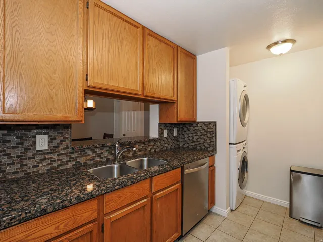 Kitchen with dishwasher, backsplash, dark stone counters, stacked washer and clothes dryer, and light tile patterned flooring