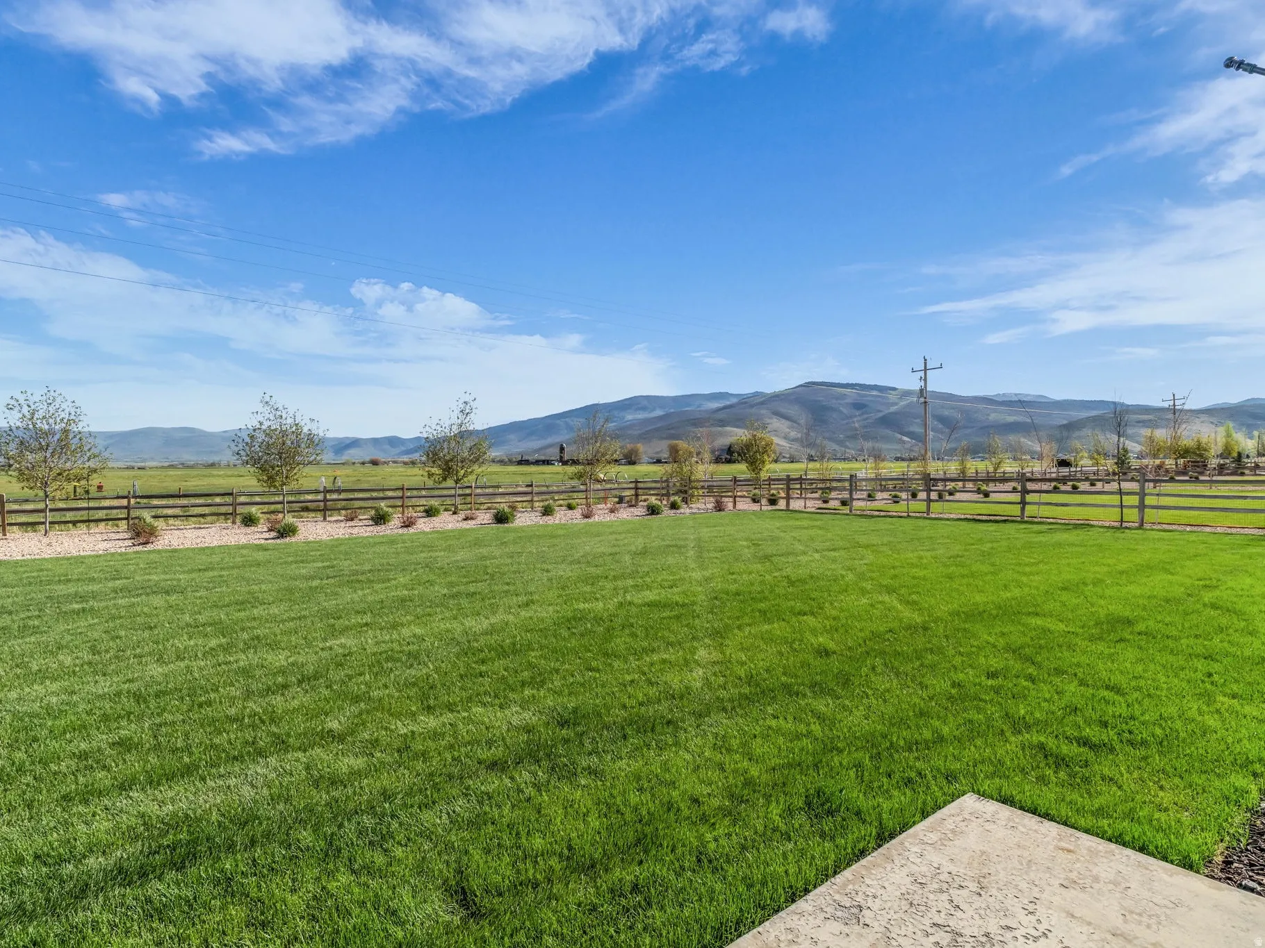Expansive green lawn with mountain backdrop, bordered by a split-rail fence, gravel landscaping, and mature trees
