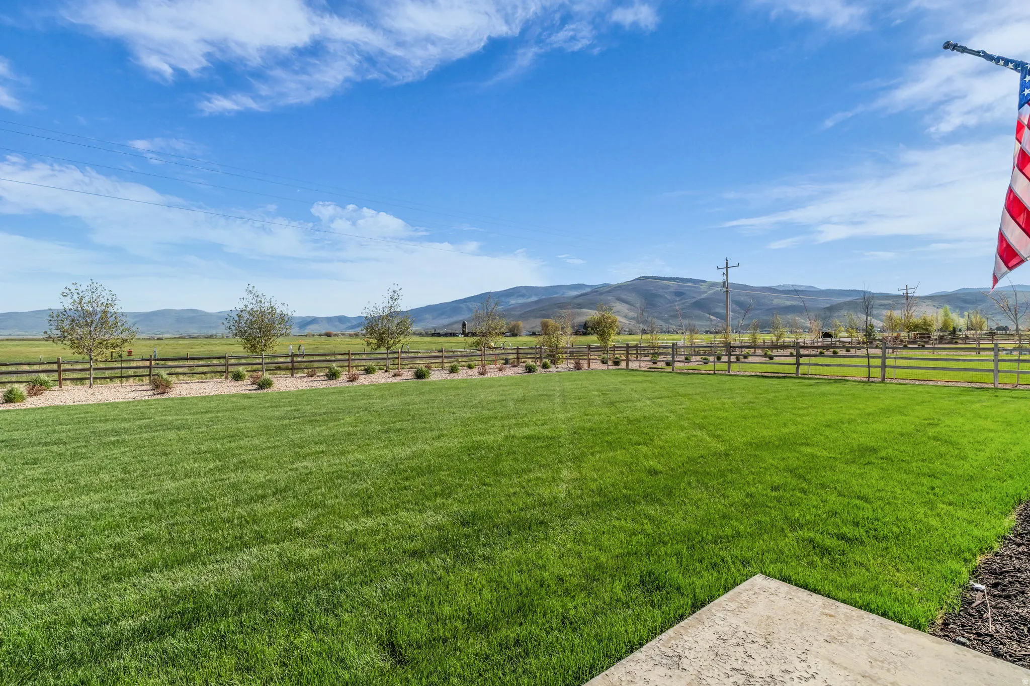 Expansive green lawn with mountain backdrop, bordered by a split-rail fence, gravel landscaping, and mature trees