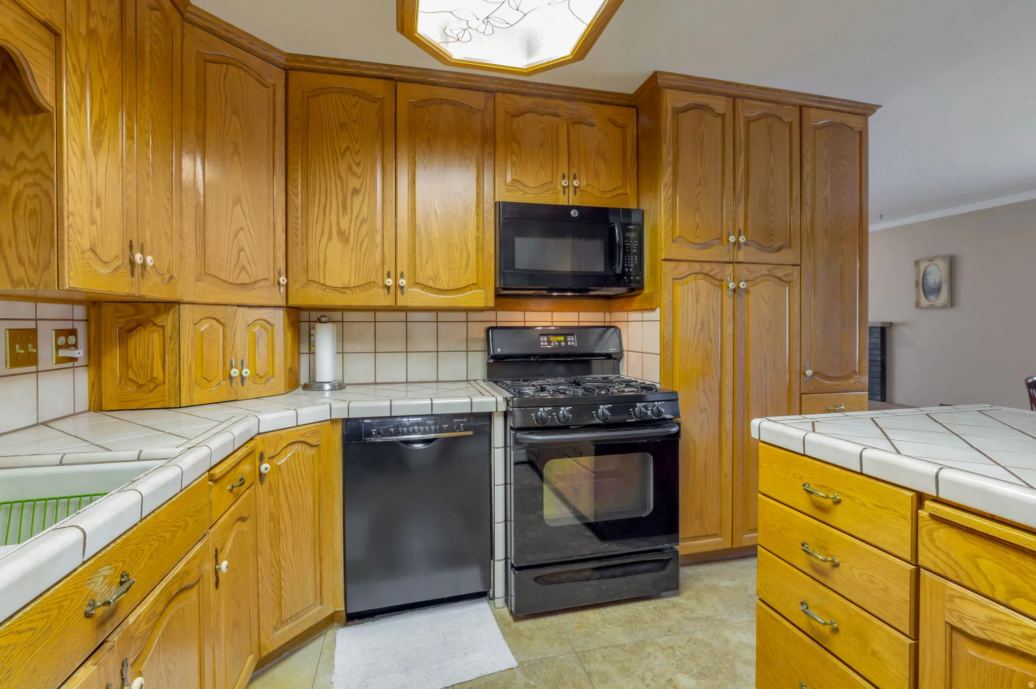 Kitchen with tile countertops, black appliances, wood finish cabinetry, ornamental molding, and tasteful backsplash
