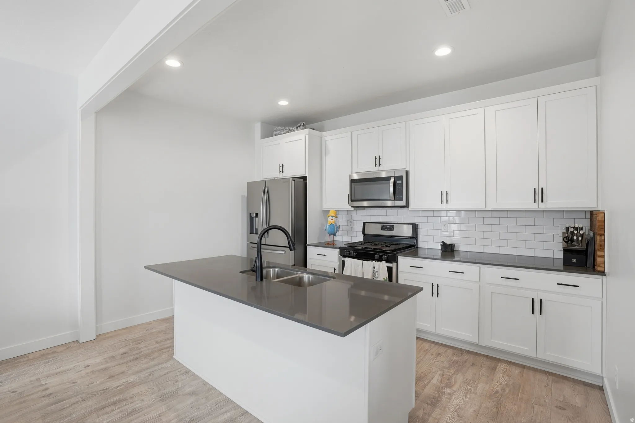 Kitchen with stainless steel appliances, light wood-type flooring, white cabinets, backsplash, and a center island with sink