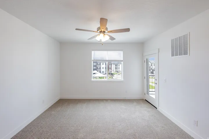 Neutral tone-on-tone carpeting, window with blinds, ceiling fan with integrated lighting, glass-paneled door, and white walls
