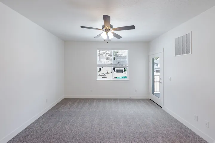 Neutral tone wall color, textured carpeting, ceiling fan with integrated lighting, window with blinds, and a glass-paneled door leading to an exterior balcony