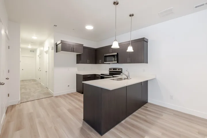 Contemporary kitchen featuring dark wood-finish cabinetry, light countertops, stainless steel appliances, a kitchen island with a sink, and pendant lighting