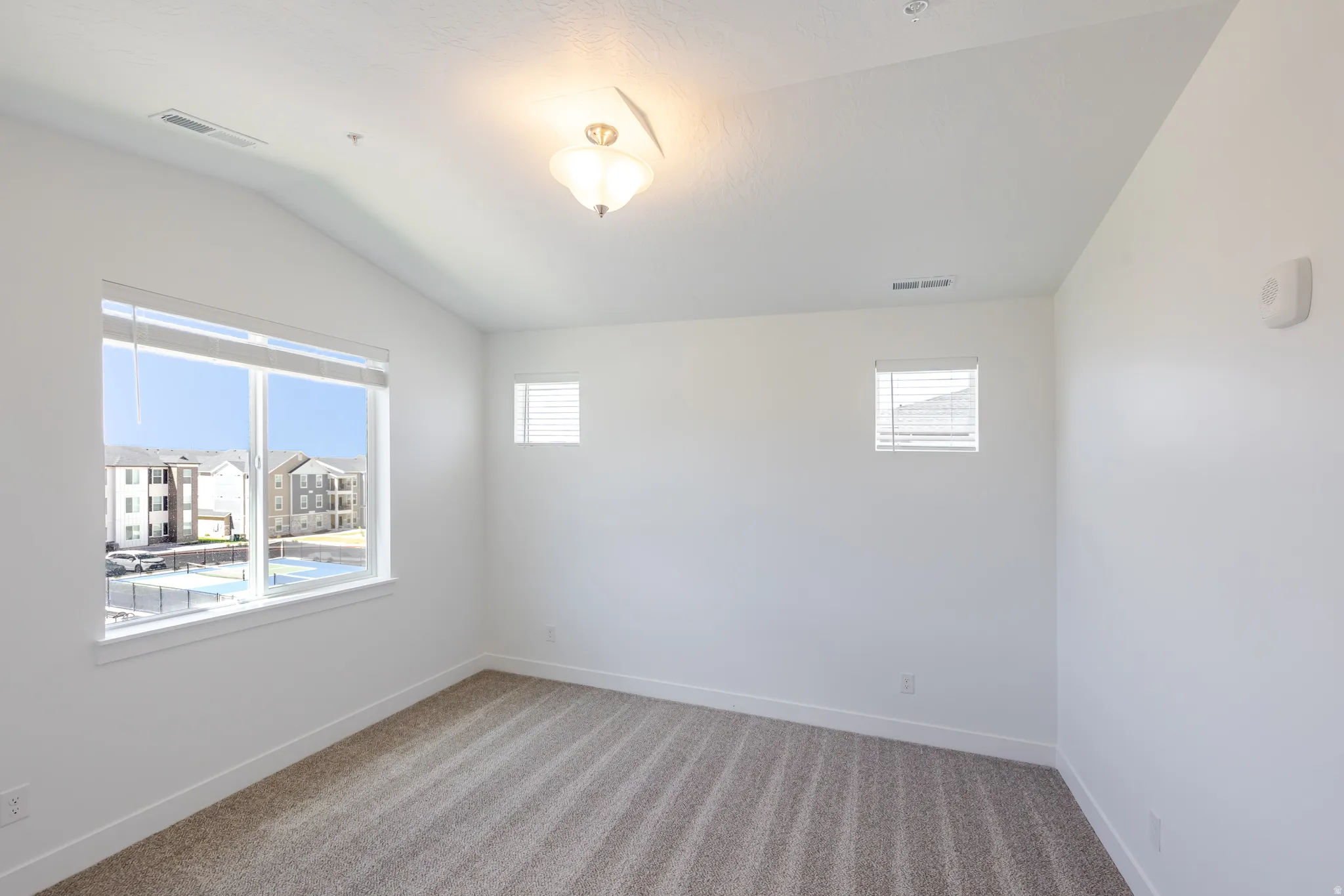 Vaulted ceiling with an attached light fixture, large windows with blinds, and neutral carpet flooring
