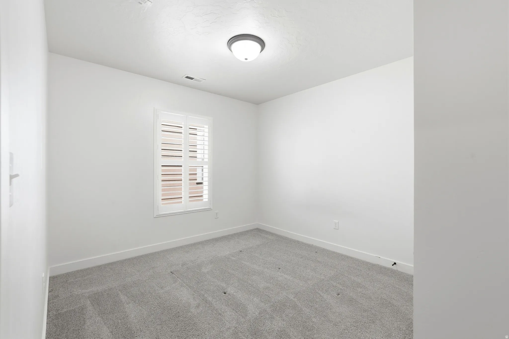 Bright room featuring white walls, gray carpet, a single window with white plantation shutters, and a flush-mount ceiling light fixture