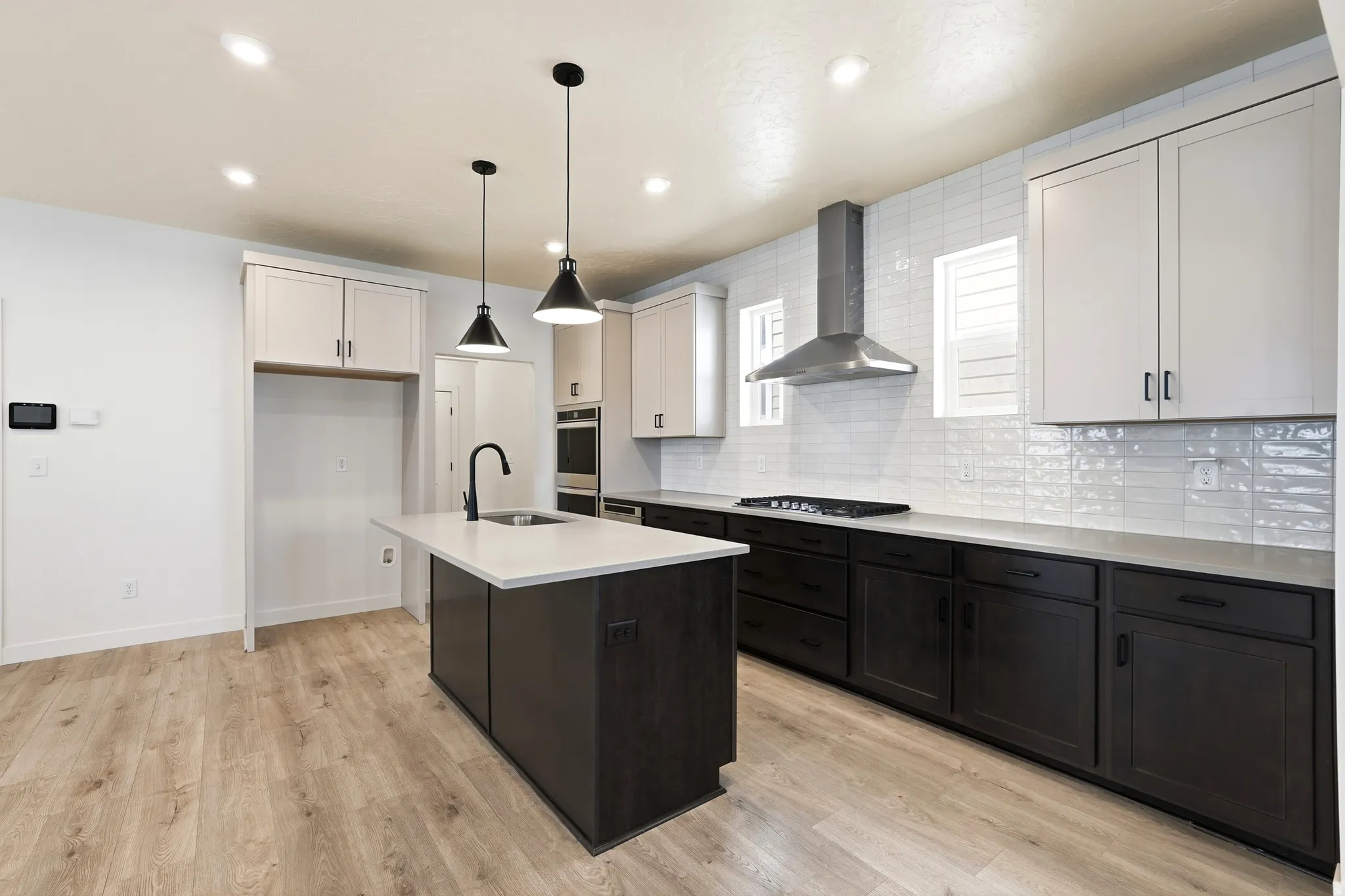 Modern kitchen featuring a central island with a sink and pendant lighting, dual-tone cabinetry, light wood-finish flooring, a stainless steel range hood, and white subway tile backsplash