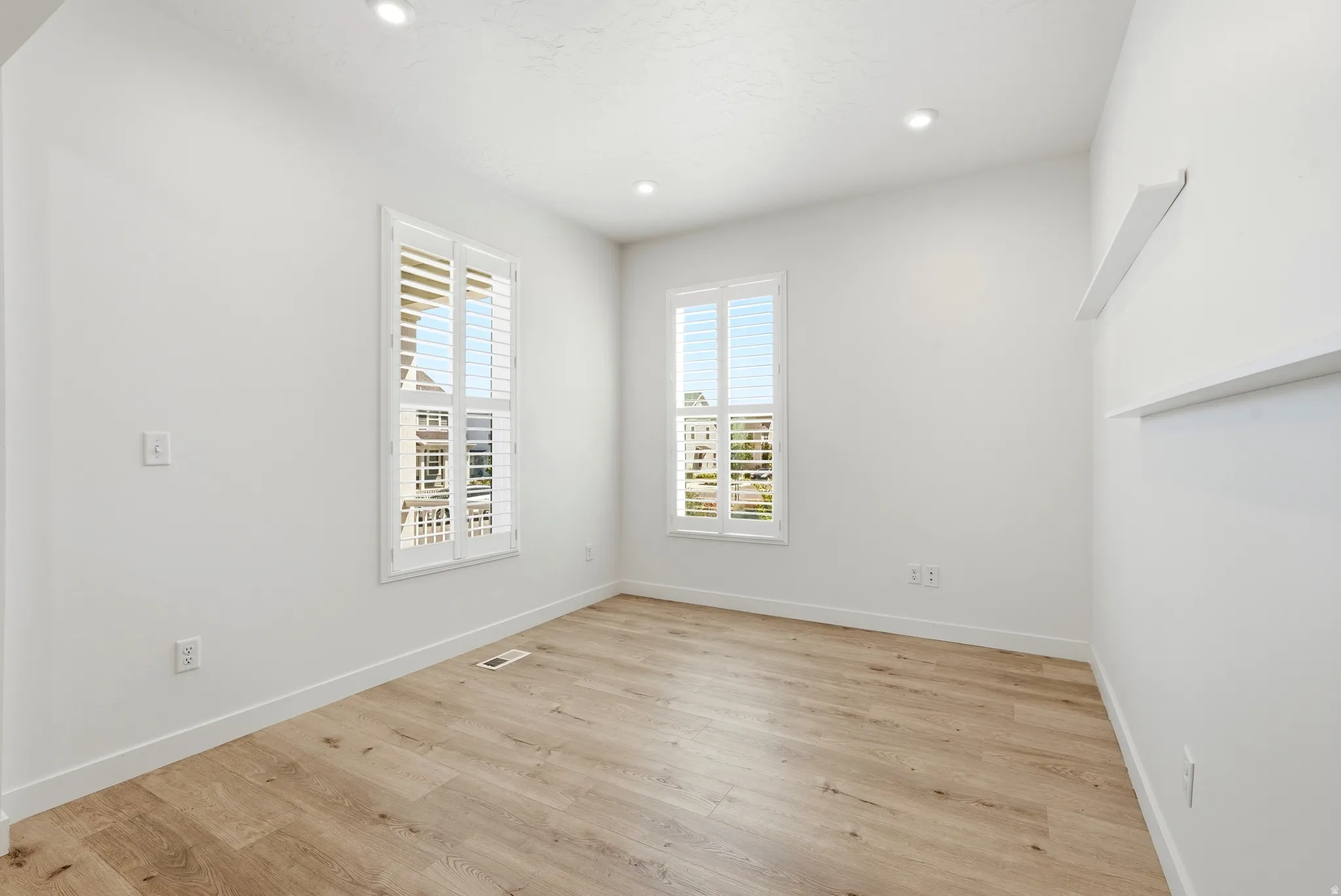 Bright room featuring two windows with plantation shutters, recessed lighting, wood-finish flooring, and built-in wall shelving