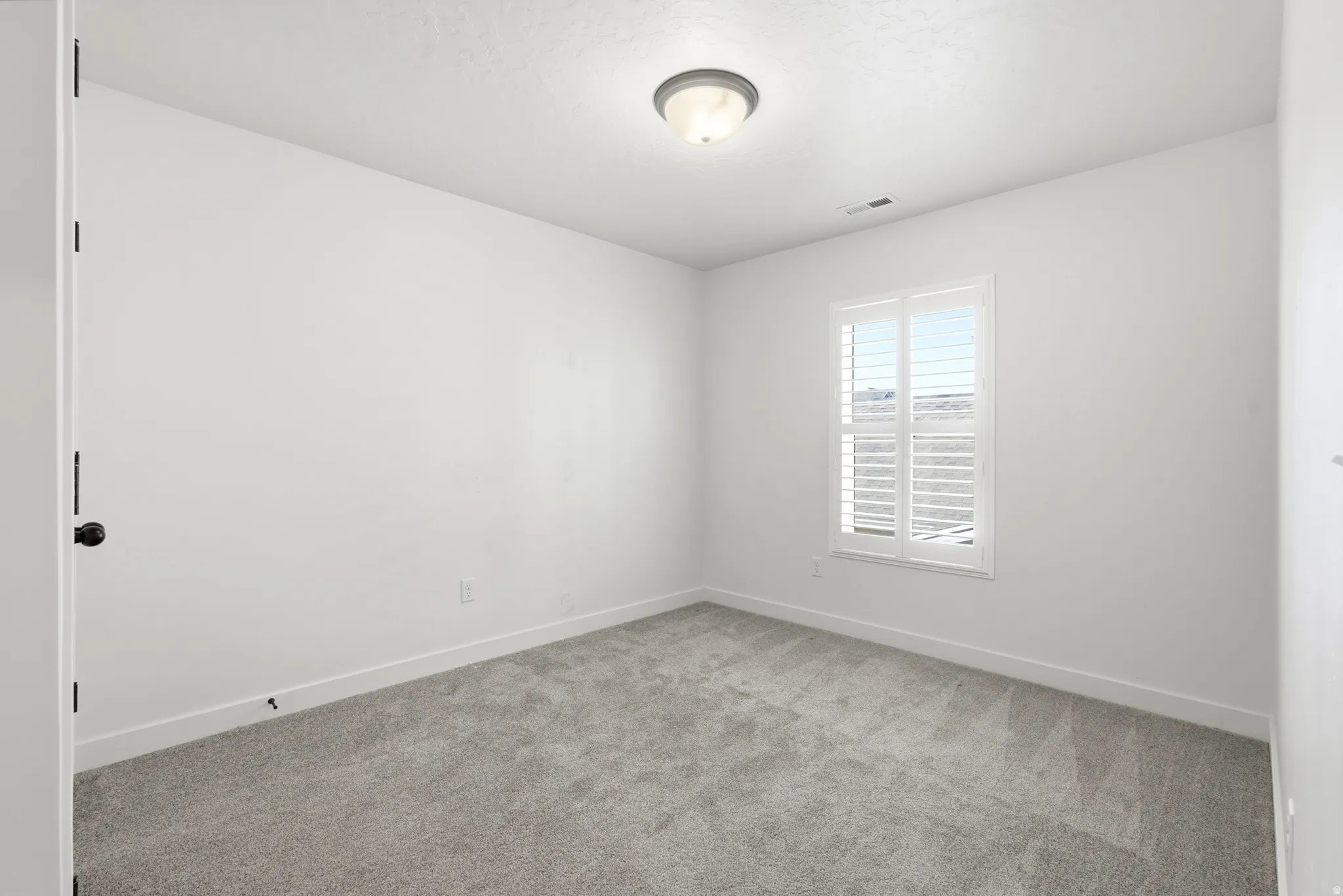 Neutral-toned room featuring a single window with white plantation shutters, light gray carpet, white walls, and a ceiling-mounted light fixture