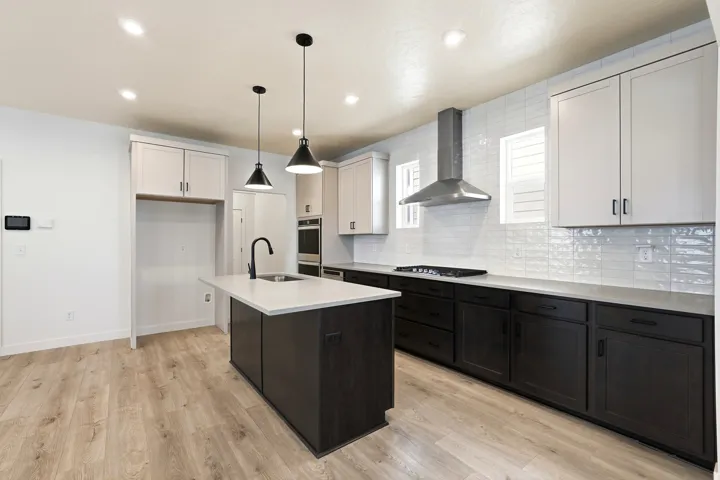 Modern kitchen featuring a central island with a sink and pendant lighting, dual-tone cabinetry, light wood-finish flooring, a stainless steel range hood, and white subway tile backsplash