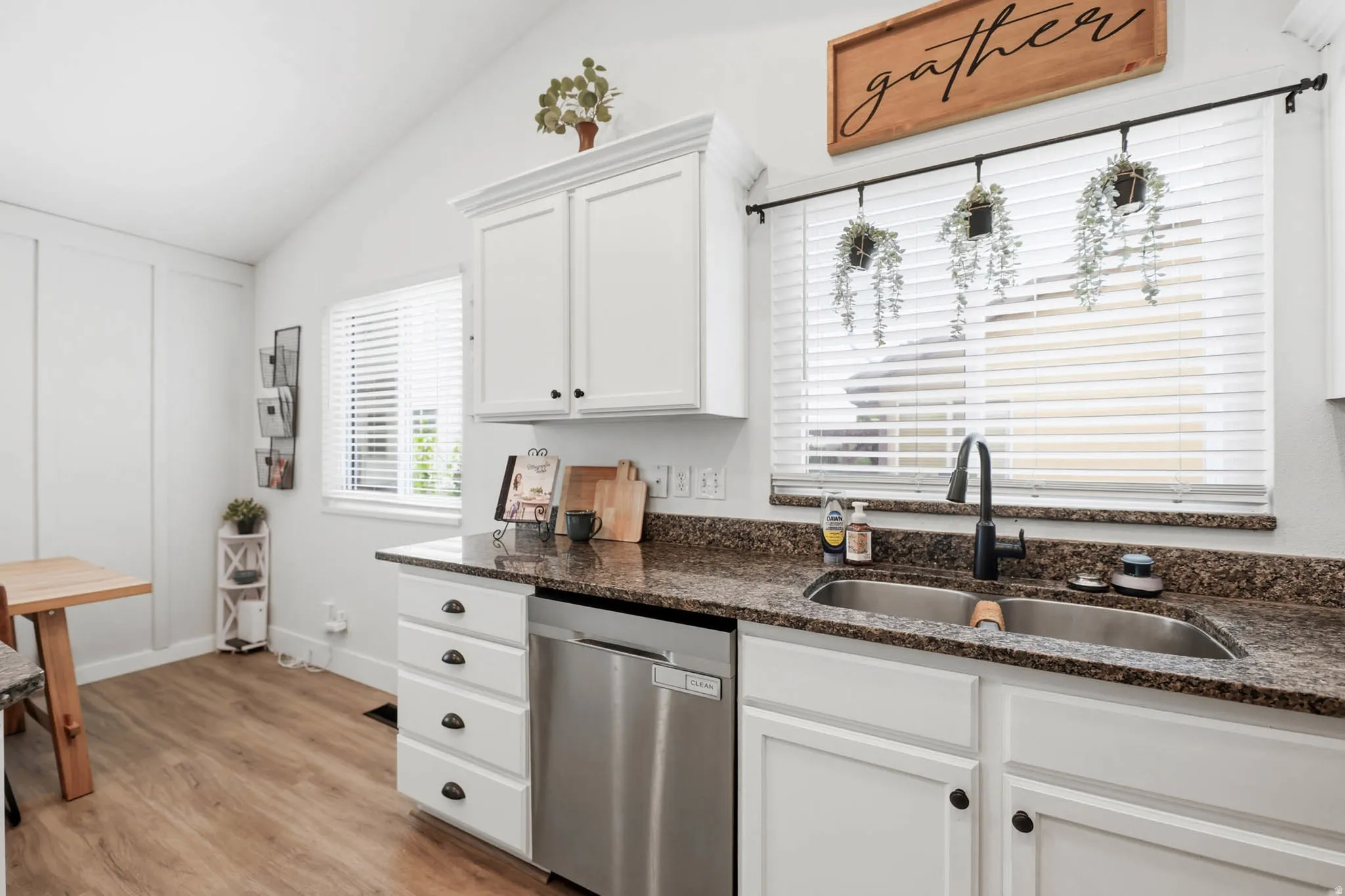 Kitchen featuring white cabinets, stainless steel dishwasher, light wood finished floors, dark stone counters, and lofted ceiling