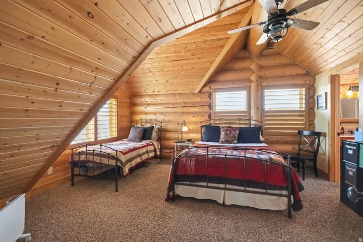 Carpeted bedroom with a vaulted wood ceiling, rustic walls, multiple windows, and a ceiling fan