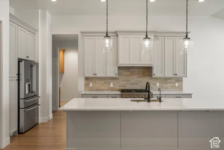 Open-concept kitchen featuring an island with a built-in sink, stainless steel appliances, light-toned cabinetry, wood-finish flooring, and pendant lighting