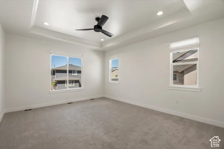 Tray ceiling with recessed lighting, ceiling fan, multiple windows with roller shades, and neutral carpeting