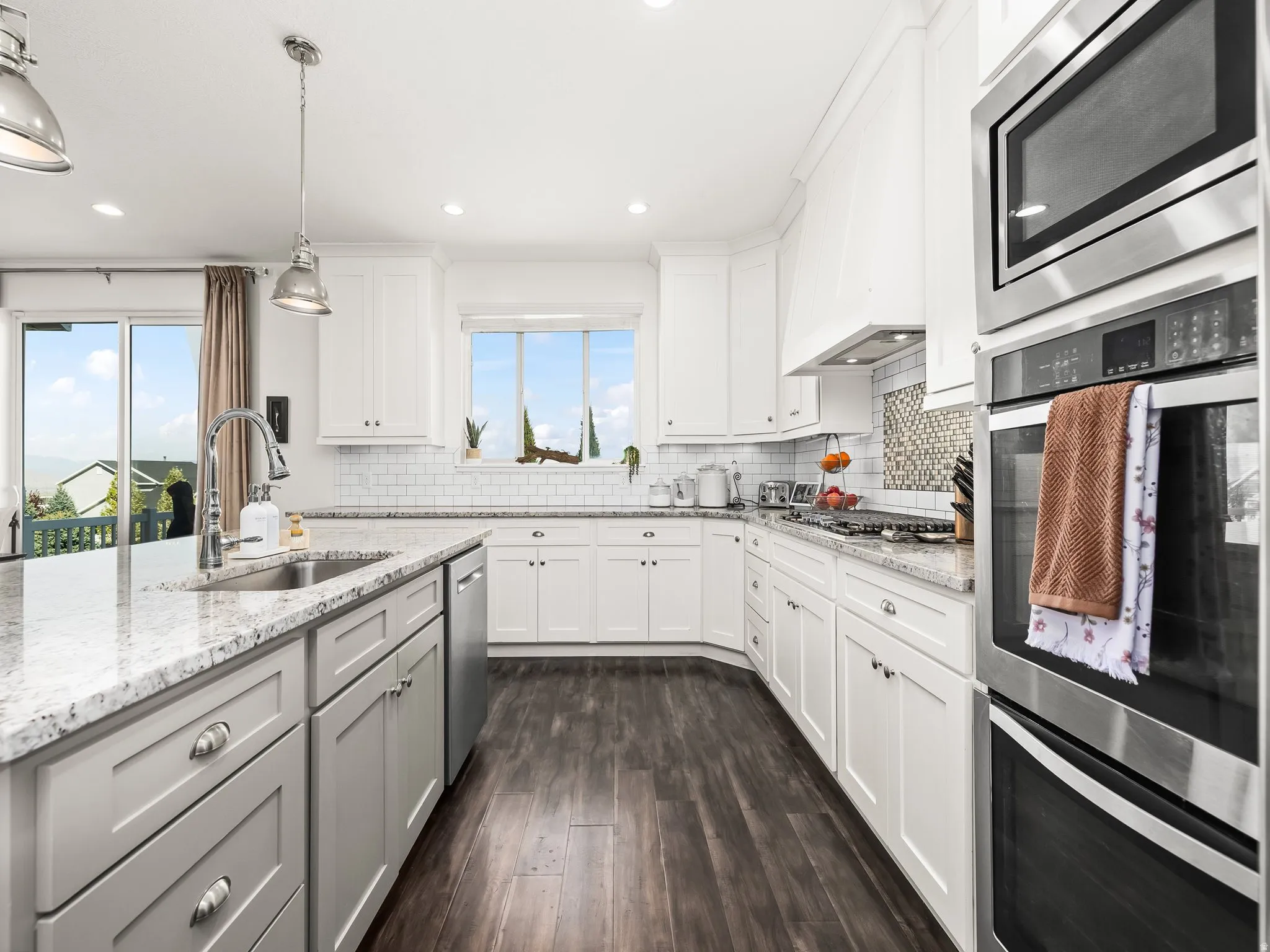 Kitchen featuring stainless steel appliances, dark wood-style flooring, light stone countertops, decorative backsplash, and hanging light fixtures