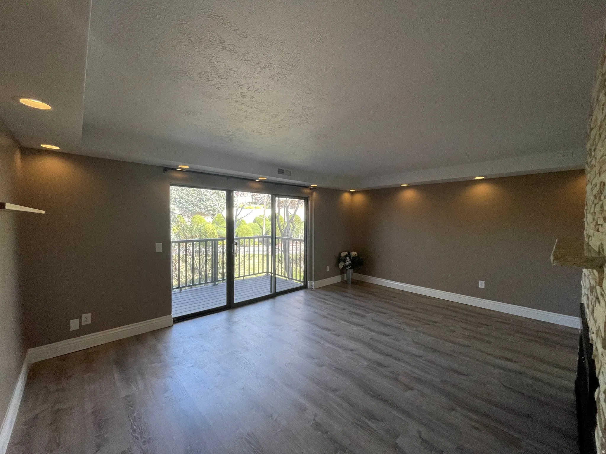 Unfurnished living room with a textured ceiling, dark wood-style floors, and recessed lighting and glass slider and windows to private deck overlooking the community lake with fountain and gorgeous mature trees.