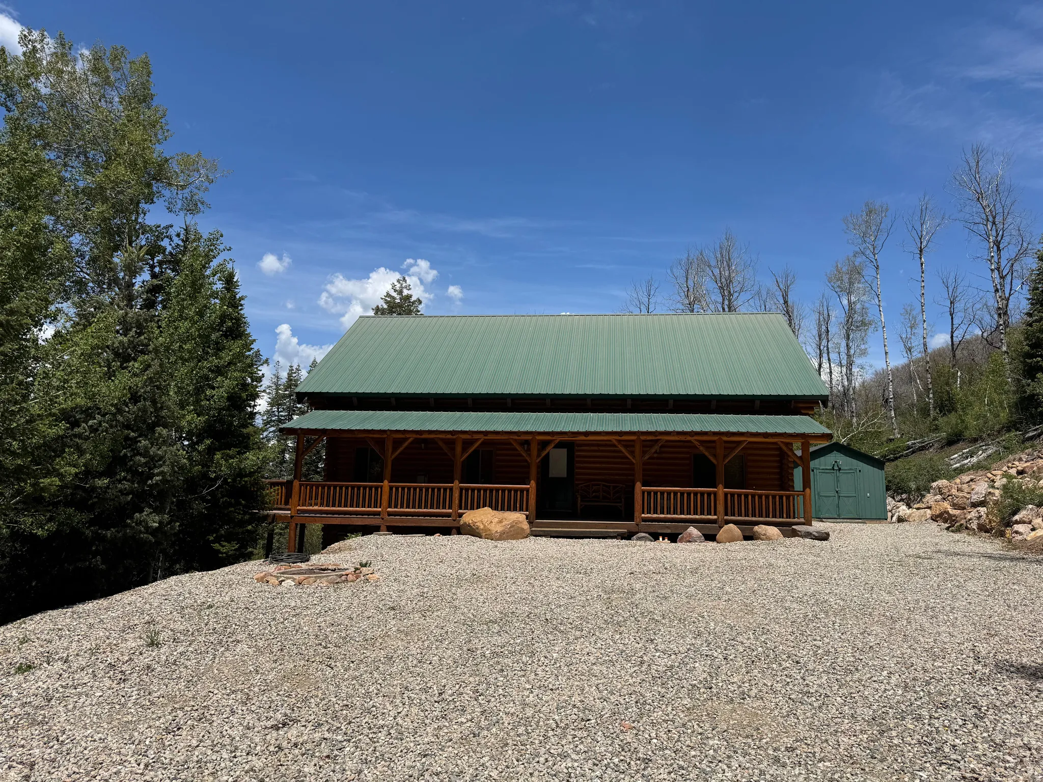 Log cabin exterior featuring a green metal roof, a full-width covered porch with wood railings, and a gravel driveway