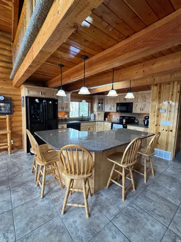 Kitchen featuring a wooden ceiling with exposed beams, black appliances, a spacious island, decorative light fixtures, and dark granite  countertops