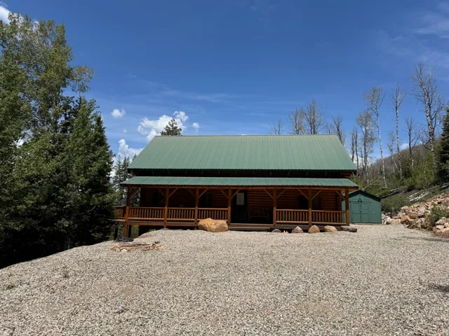 Log cabin exterior featuring a green metal roof, a full-width covered porch with wood railings, and a gravel driveway
