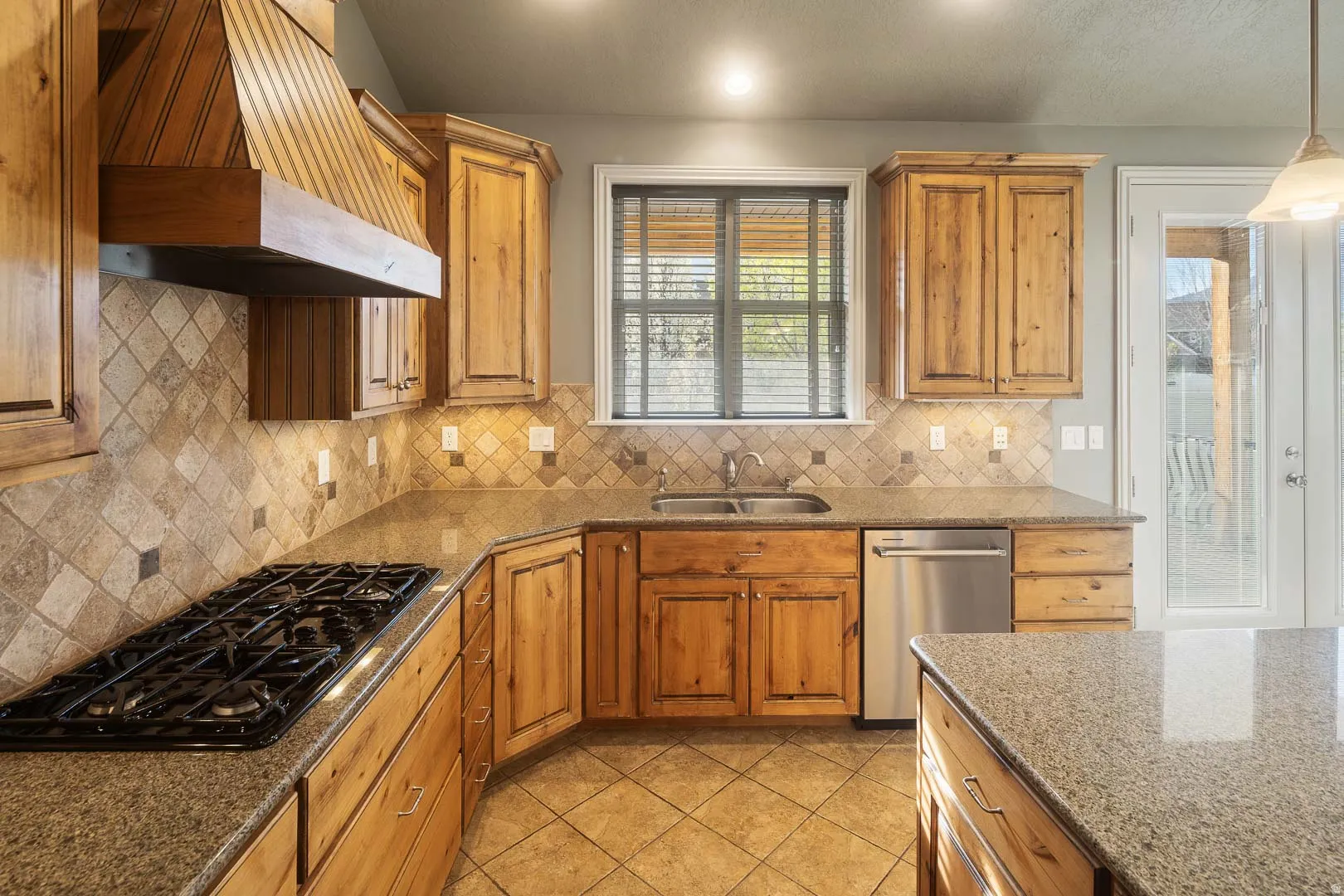 Kitchen featuring dark stone counters, dishwasher, black gas cooktop, wood finish cabinets, and light tile patterned flooring