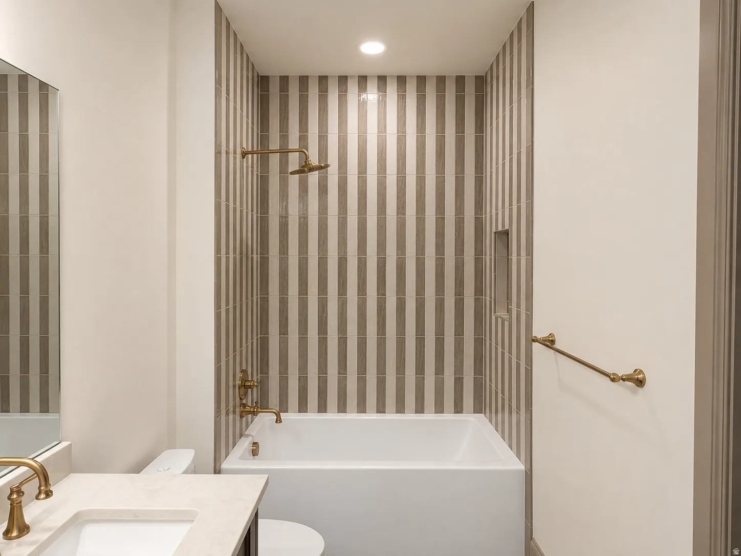 Bathtub with brass fixtures, vertically-striped tiled shower surround, recessed shower niche, and built-in vanity with single sink