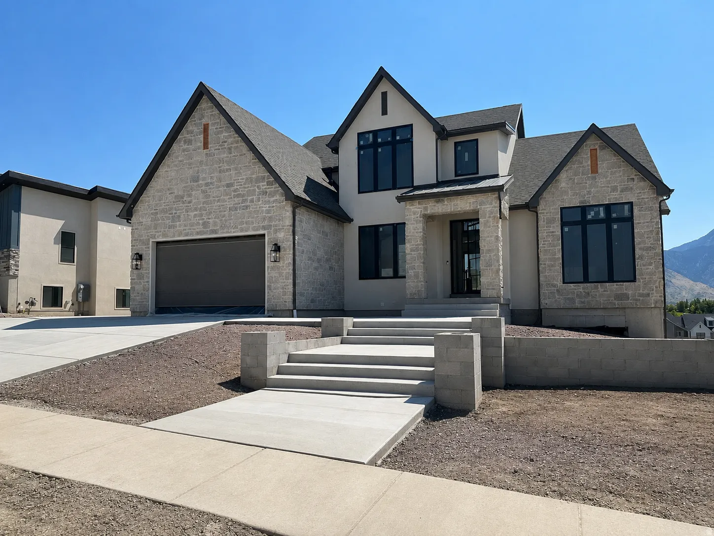 Modern architectural design featuring a prominent gabled roofline, stone veneer and stucco facade, an integrated garage with a dark door, and a multi-level concrete walkway leading to the entrance